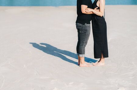 young couple a guy with a girl in black clothes are walking on the white sand at the edge of blue waterの写真素材