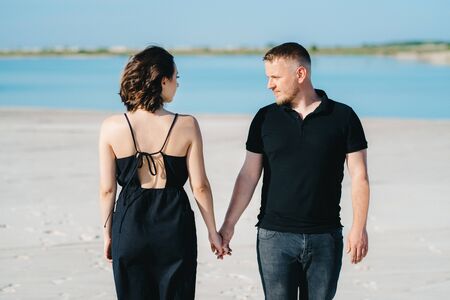 young couple a guy with a girl in black clothes are walking on the white sand at the edge of blue waterの写真素材