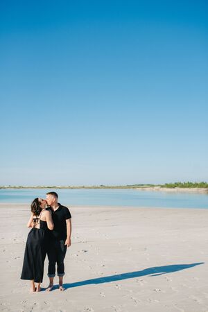 young couple a guy with a girl in black clothes are walking on the white sand at the edge of blue waterの写真素材