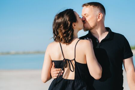 young couple a guy with a girl in black clothes are walking on the white sand at the edge of blue waterの写真素材