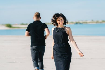 young couple a guy with a girl in black clothes are walking on the white sand at the edge of blue waterの写真素材