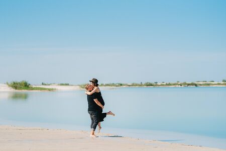 young couple a guy with a girl in black clothes are walking on the white sand at the edge of blue waterの写真素材