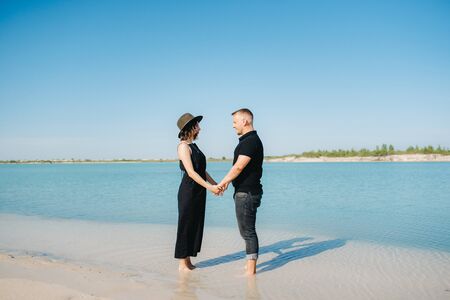 young couple a guy with a girl in black clothes are walking on the white sand at the edge of blue waterの写真素材