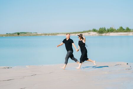 young couple a guy with a girl in black clothes are walking on the white sand at the edge of blue waterの写真素材