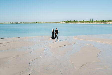 young couple a guy with a girl in black clothes are walking on the white sand at the edge of blue waterの写真素材
