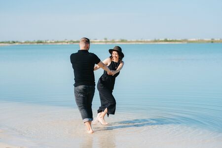 young couple a guy with a girl in black clothes are walking on the white sand at the edge of blue waterの写真素材