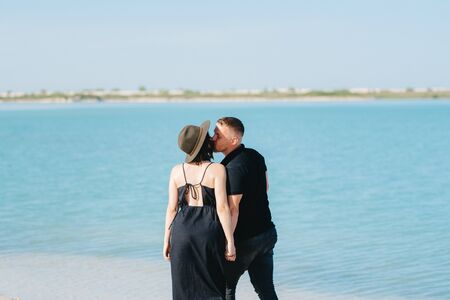 young couple a guy with a girl in black clothes are walking on the white sand at the edge of blue waterの写真素材