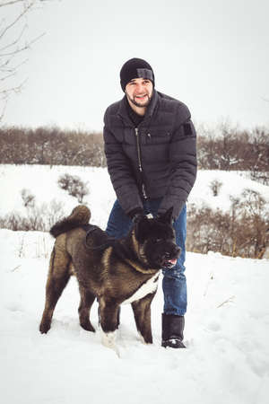 A man in a jacket and a knitted hat walks through a snowy forest with an American Akita dogの写真素材