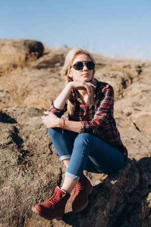 blonde girl with pigtails in a shirt, jeans, red shoes and black glasses on granite rocksの写真素材
