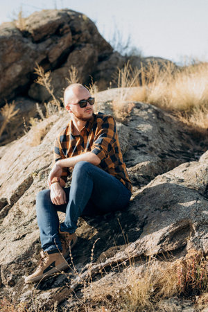 bald guy with a beard in jeans warm shirt and trekking shoes on granite rocksの写真素材
