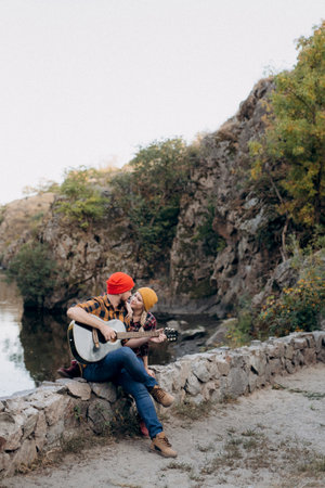 a guy in a bright hat plays the guitar with a girl against a background of granite rocks and a riverの写真素材