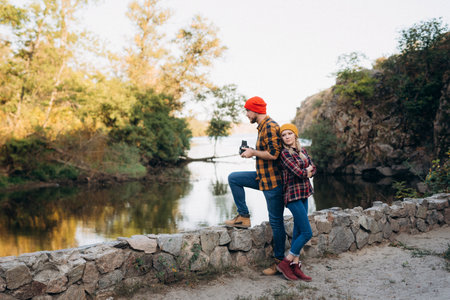 Bald guy with a beard and a blonde girl in bright hats on the background of the river are taking pictures with an old cameraの写真素材