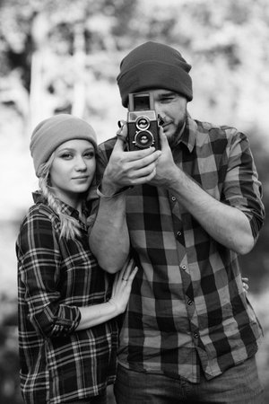 Bald guy with a beard and a blonde girl in bright hats on the background of the river are taking pictures with an old cameraの写真素材