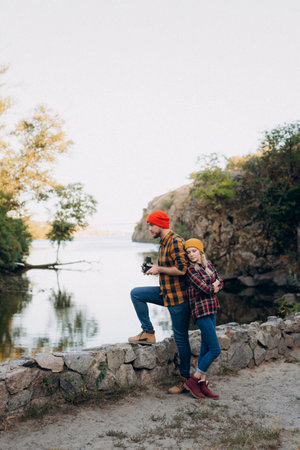 Bald guy with a beard and a blonde girl in bright hats on the background of the river are taking pictures with an old cameraの写真素材
