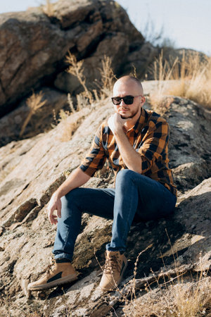 bald guy with a beard in jeans warm shirt and trekking shoes on granite rocksの写真素材