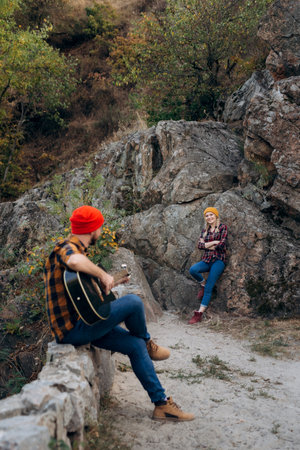 a guy in a bright hat plays the guitar with a girl against a background of granite rocks and a riverの写真素材