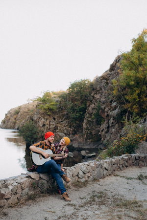 a guy in a bright hat plays the guitar with a girl against a background of granite rocks and a riverの写真素材