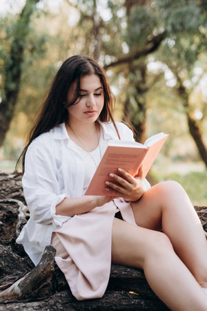 young girl in white clothes sits on a tree in the forest and reads a bookの写真素材