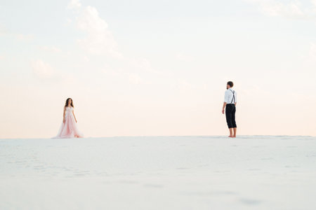 young couple a guy in black breeches and a girl in a pink dress are walking along the white sand of the desertの写真素材