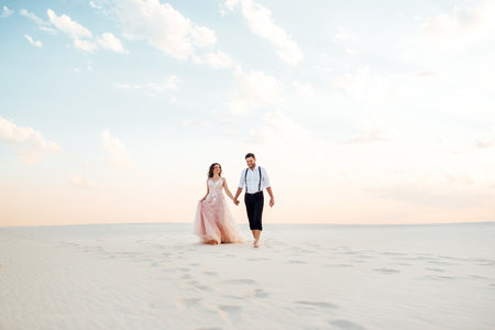 young couple a guy in black breeches and a girl in a pink dress are walking along the white sand of the desertの写真素材
