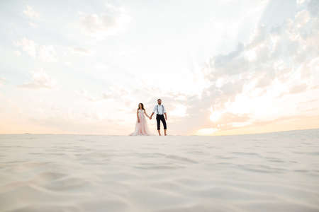 young couple a guy in black breeches and a girl in a pink dress are walking along the white sand of the desertの写真素材