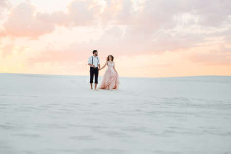 young couple a guy in black breeches and a girl in a pink dress are walking along the white sand of the desertの写真素材