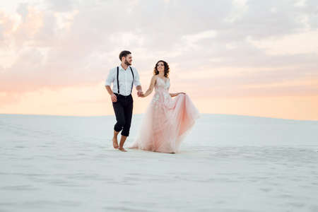 young couple a guy in black breeches and a girl in a pink dress are walking along the white sand of the desertの写真素材
