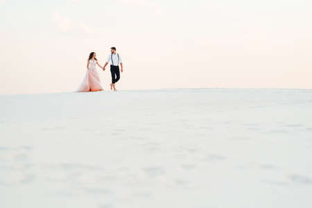 young couple a guy in black breeches and a girl in a pink dress are walking along the white sand of the desertの写真素材
