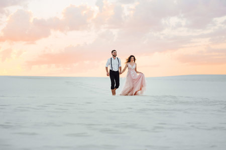 young couple a guy in black breeches and a girl in a pink dress are walking along the white sand of the desertの写真素材