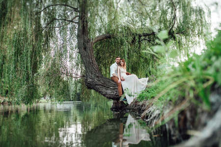 a guy and a girl are sitting together on a crooked tree on the steep bank of a wild riverの写真素材