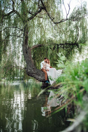 a guy and a girl are sitting together on a crooked tree on the steep bank of a wild riverの写真素材
