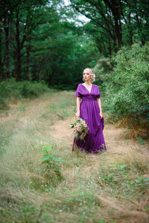 Girl model blonde in a lilac dress with a bouquet with a green forestの写真素材