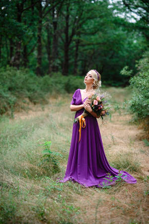 Girl model blonde in a lilac dress with a bouquet with a green forestの写真素材