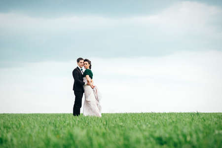 the groom in a brown suit and the bride in an ivory-colored dress on a green field receding into the distance against the skyの写真素材