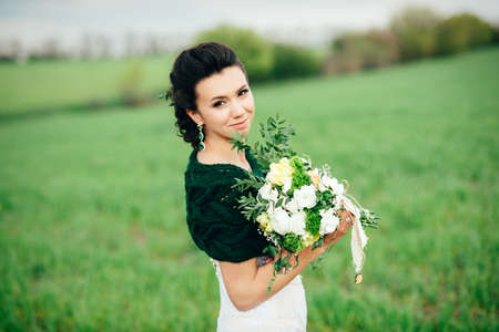 bride with a bouquet in an ivory dress and a knitted shawl on a green fieldの写真素材