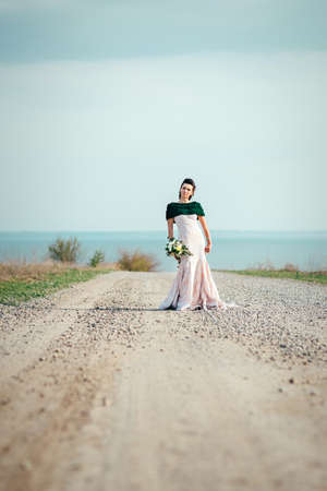 bride with a bouquet of flowers in an ivory dress and a knitted shawl on the road stretching into the distanceの写真素材