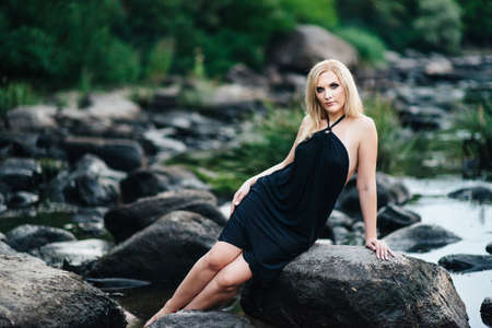 blonde girl in a black dress with blue eyes on the stone coast of the riverの写真素材