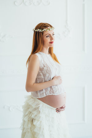 red-haired pregnant young girl in a white dress near the white wall of a large bright roomの写真素材