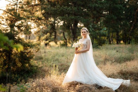 bride blonde girl with a bouquet in the forest in the sunset lightの写真素材