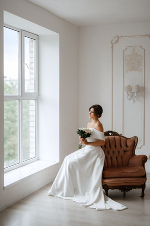 brunette bride in a tight wedding dress in a bright studio with a bouquet on an armchairの写真素材