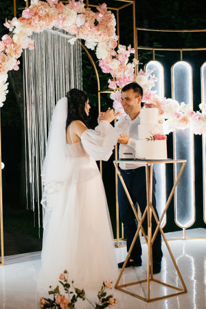 newlyweds happily cut the wedding cake on the wedding arch, laugh and taste the wedding cakeの写真素材