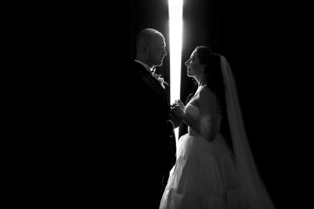 A black-haired bride in a white dress and a groom in a blue suit with a bow tie in a dark hotel roomの写真素材