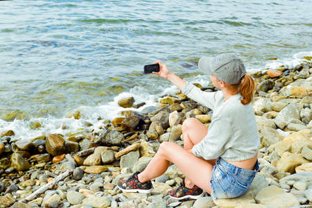 young woman in gray hoodie and denim shorts with a smartphone on the rocky shoreの写真素材