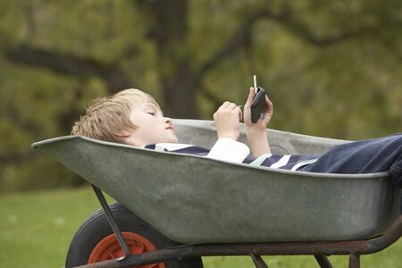 Young Boy Laying Wheelbarrow Using Smart Mobile Phoneの写真素材