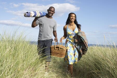 Young Couple Carrying Picnic Basket And Windbreak Walking Through Dunesの写真素材