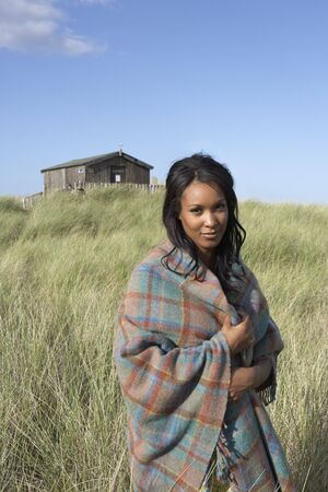 Young Woman Standing On Beach Wrapped In Blanket With Beach Hut In Distanceの写真素材