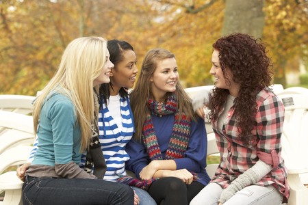 Group Of Four Teenage Girls Sitting And Chatting On Bench In Autumn Parkの写真素材