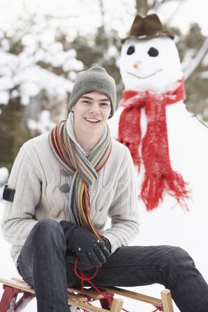Teenage Boy With Sledge Next To Snowmanの写真素材