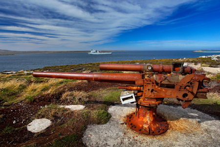 Falklands War, rocky coast with old rusty cannon. Corroded artillery gun from Falklands Conflict in nature habitat. Blue sky landscape, Falkland Islands. Military ship on sea. Memorial war monument.の写真素材