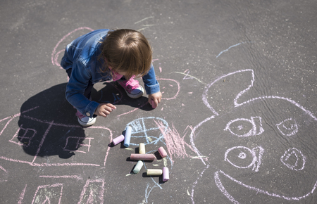 Little beautiful girl in a jeans jacket draws with colored chalks on the playgroundの写真素材